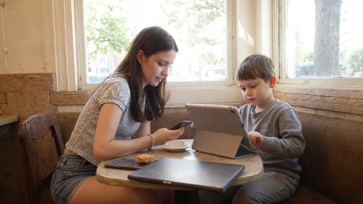A young woman checks her phone at a cafe table by the window while a little boy beside her taps on a tablet propped on a stand.