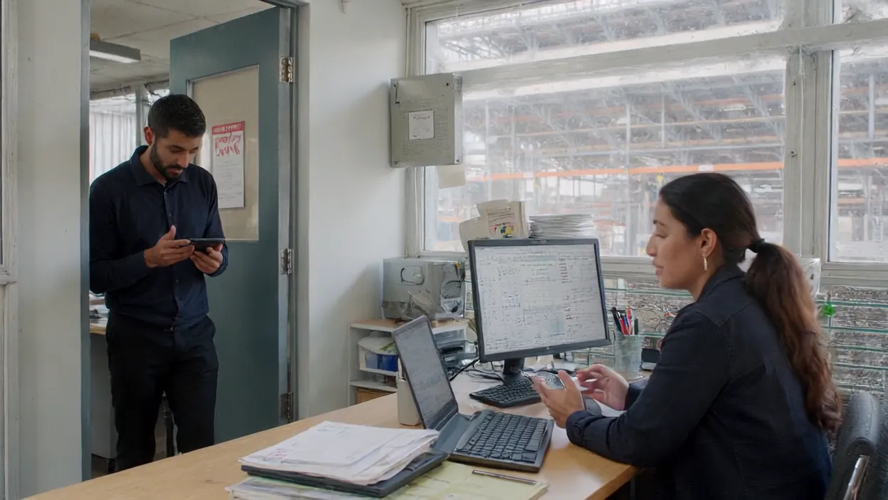 An IT staffer checks his phone in a doorway while an operations manager works at her computer.