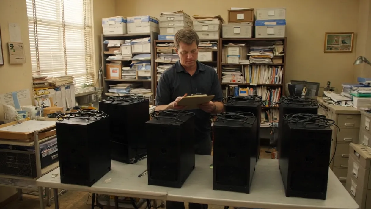 A technician with a clipboard checks a row of black desktop computers on a table in a back office.